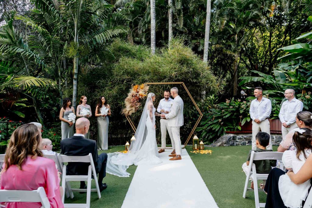 the ring bearer presenting the ring in a modern seamless inclusive way, wfacing the wedding guests, so everyone can see the moment. Framwd by a gold hexagonal arbour stiid between the bride and groom as he hands them their wedding rings