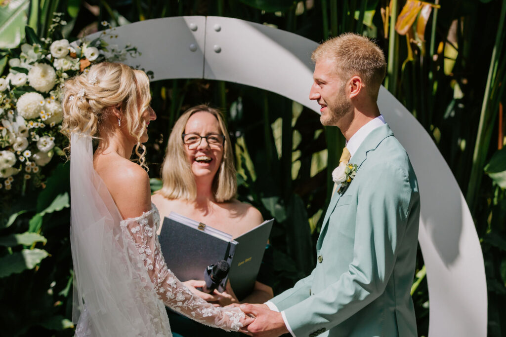 Celebrant Cara delivering her signature happy, fun, enjoyable ceremony with the Celebrant and bride and groom laughing along during the ceremony framed by the white circular wedding arch